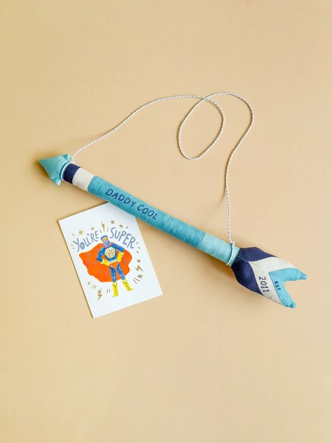 A handmade linen arrow wall hanging in shades of blue and navy, embroidered with the words “DADDY COOL” along the shaft and “2011” on the fletching. A white and grey cord is attached for hanging.