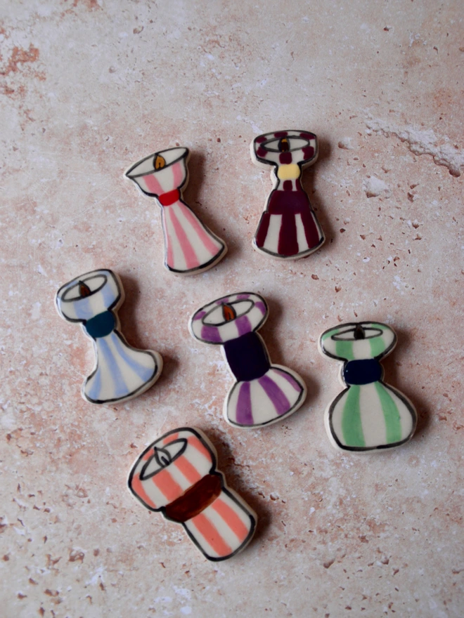 Handmade ceramic magnets shaped like candlesticks, featuring colourful stripe patterns in red, pink, green, blue and purple, arranged on a neutral textured background.