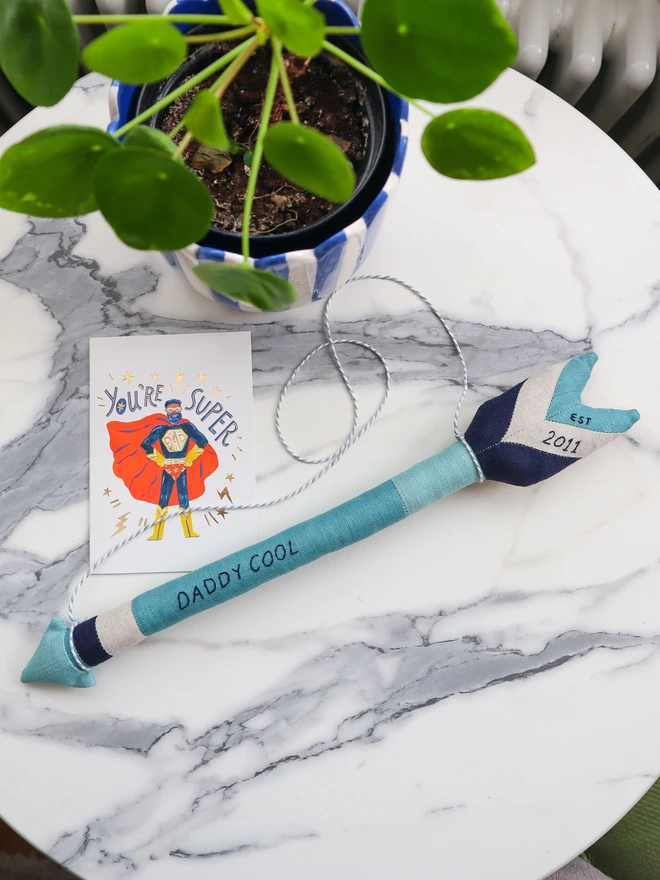 A handmade linen arrow wall hanging in shades of blue and navy, embroidered with the words “DADDY COOL” along the shaft and “2011” on the fletching. A white and grey cord is attached for hanging.