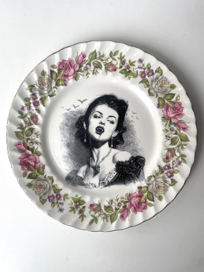A vampire with bats behind her, the image is victorian in black and white and is printed directly onto an ornate vintage plate