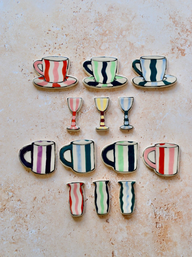 Collection of handmade ceramic miniatures featuring striped cups, saucers, goblets, mugs, and vases in various colorways, arranged neatly on a warm neutral textured background.