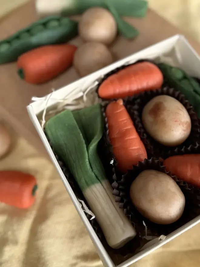 Veggilicious Marzipan Box, a marzipan vegetable box on a wooden chopping board on a yellow table cloth. In the box are marzipan peas, spring onions, potatoes and carrots. 