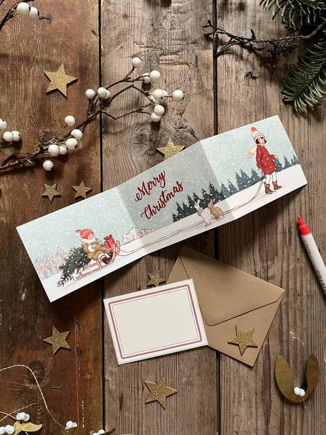 a vintage style illustrated concertina Christmas card featuring a vintgae story book style illustration of a little girl in red coat pulling a toddler on a sledge with a Christmas tree