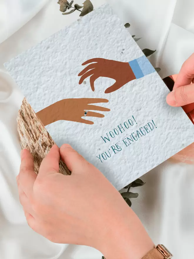 Seeded Paper Greeting Card featuring an illustration of a hand placing a ring on another hand with ‘WooHoo You’re Engaged’ underneath being placed onto a white cloth