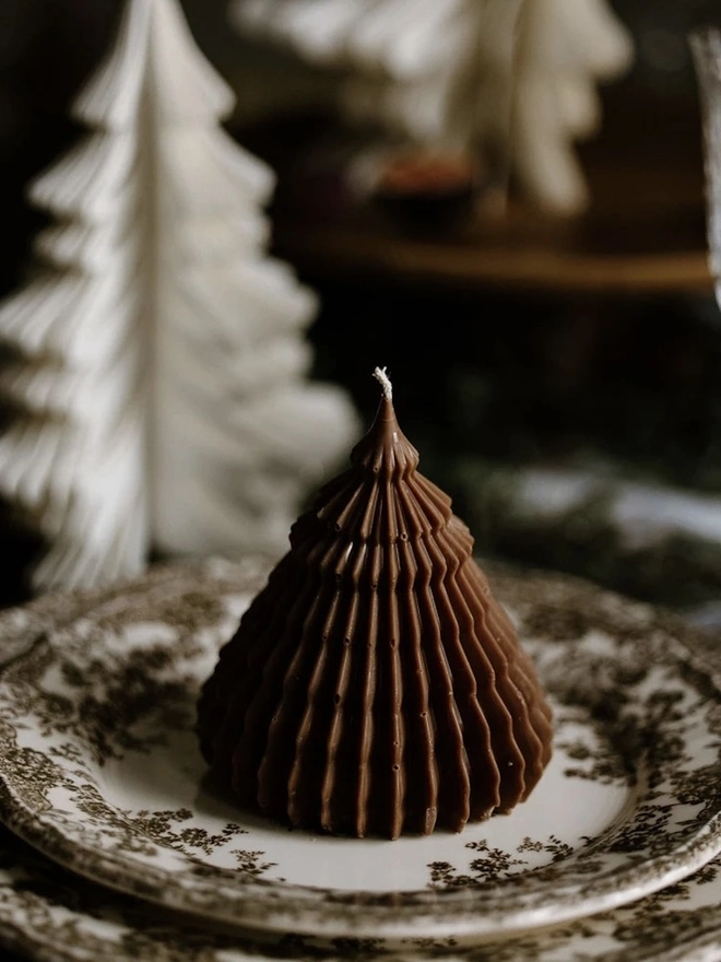 Brown Origami Christmas Tree Warm Candles (Set Of 2). Close-up of a chocolate brown cone tree candle with fine fluted ridges, set on a vintage floral transfer-print plate with a white paper tree blurred behind. The intricate sculptural texture makes it a striking festive place-setting decoration.