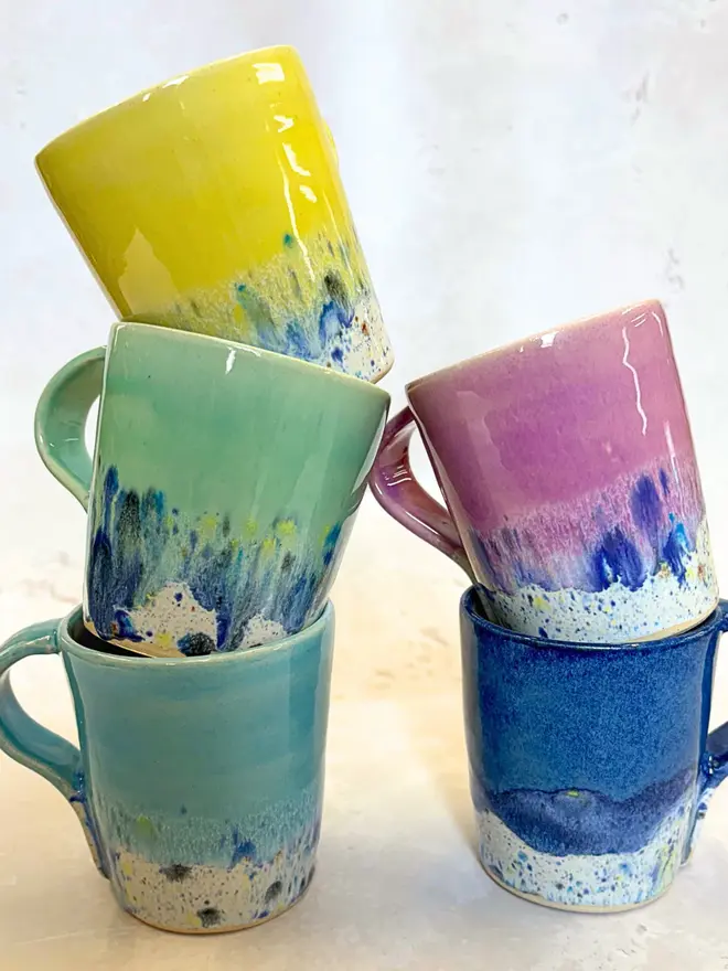 Shoreline Standard Mug, a group of colourful mugs placed on a stone surface stacked on top of each other. 