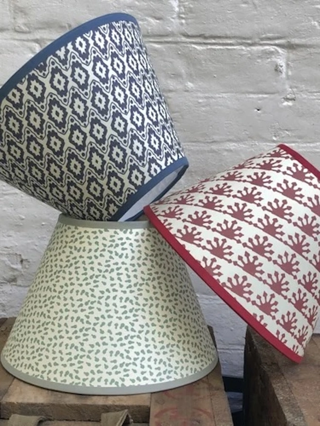 Paper Empire Lampshade. Three block-printed paper lampshades stacked against a white brick wall in navy ikat, sage scattered leaf and red sunburst designs. Each tapered shade is finished with a coordinating trim for a bold, handmade look.