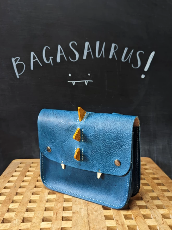 Front view of blue, hand- dyed leather 'Bagasaurus' backpack with yellow spikes and pointed white fangs.