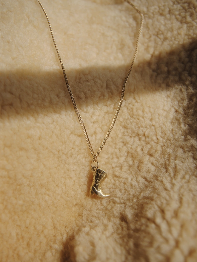 Image of hand carved cowboy boot pendant, cast in gold toned recycled brass, hanging on a 9 carat gold chain. The background of the image is sheepskin fleece fabric.