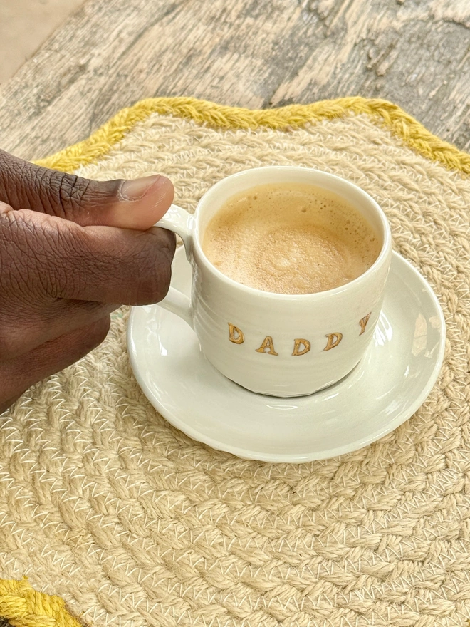 Espresso cup and saucer with DADDY emlazoned on the side with gold lustre detailing. Hand is reaching for the filled cup that is sat on a wooden farmhouse style table