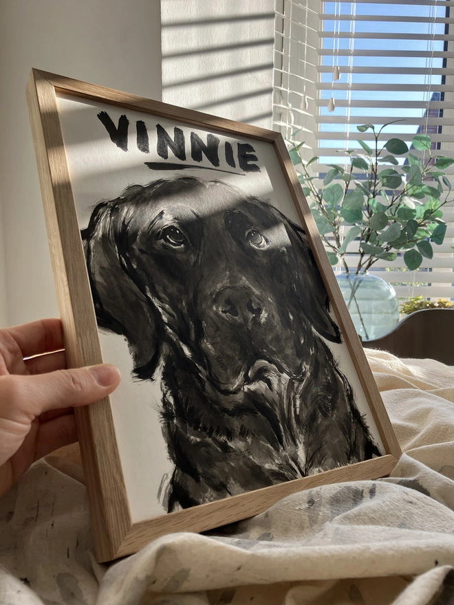 A hand holding a personalised dog portrait of a black labrador by artist 'Dogs by Jeff'