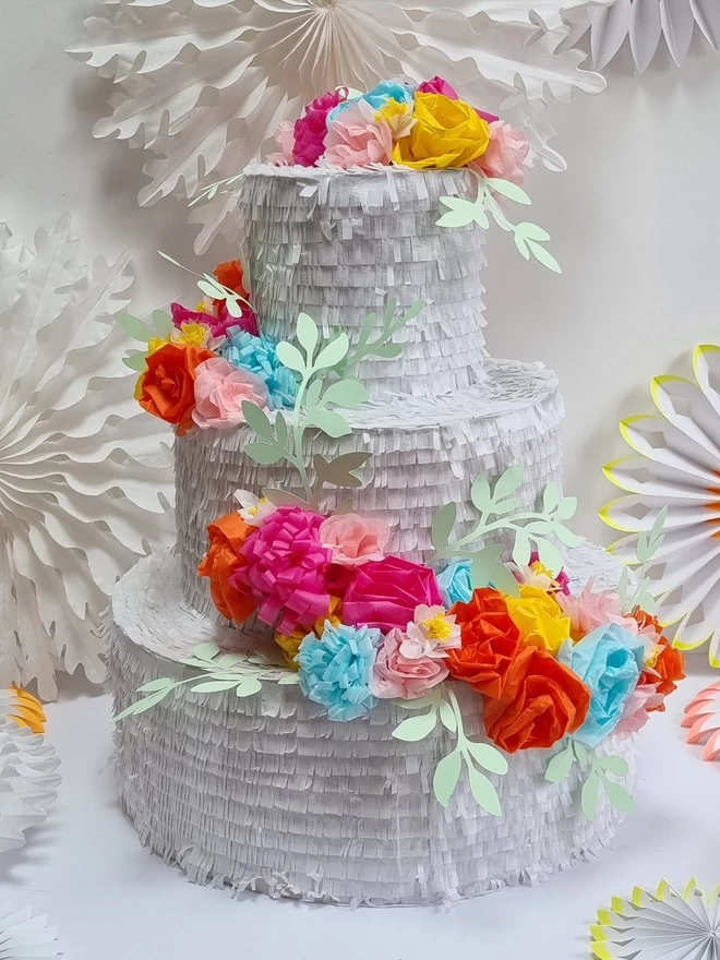 bright coloured floral wedding cake pinata on a white background surrounded by white paper decorations