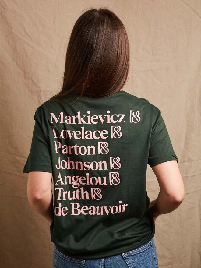 Model is showing the back of the forest green feminist icon t-shirt. The names of our chosen icons, Markievicz, Lovelace, Parton, Johnson, Angelou, Truth, and De Beauvoir printed in pale pink 