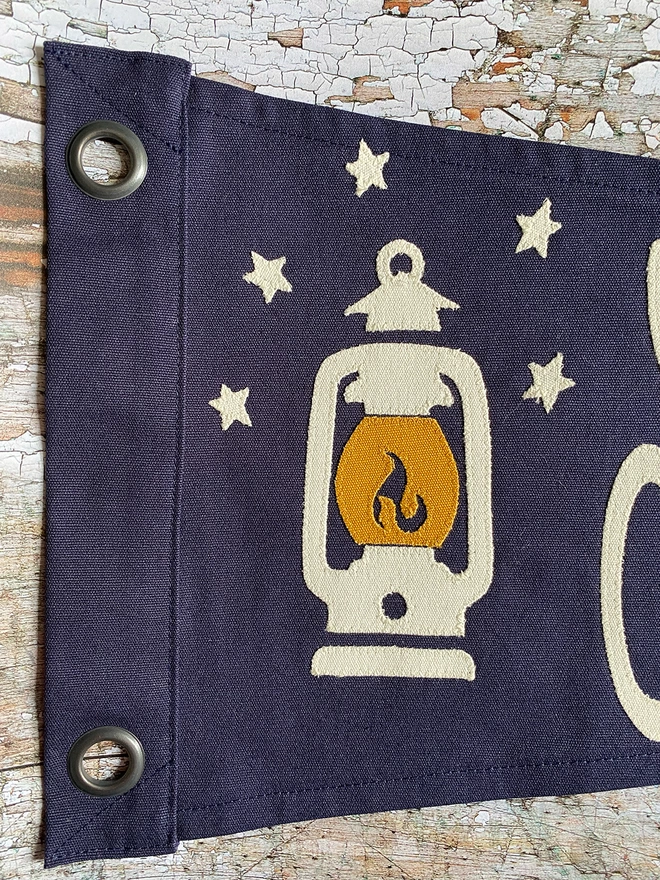 Detail of a navy canvas 'Seek Adventure' pennant flag showing the hurricane lantern in ivory and mustard canvas with some ivory canvas stars.