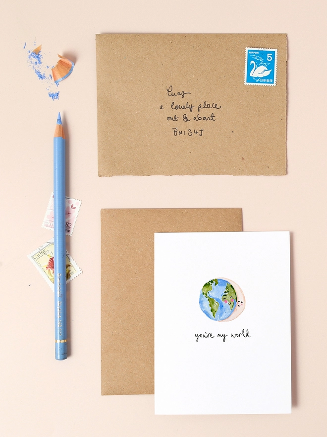 An image of a mini, rectangular white greetings card with an illustration of a smiling earth hugging a smiling crescent moon with a hand written 'Big love' message in black ink seen under the central illustration. The card is seen on top of a brown rectangular kraft envelope.