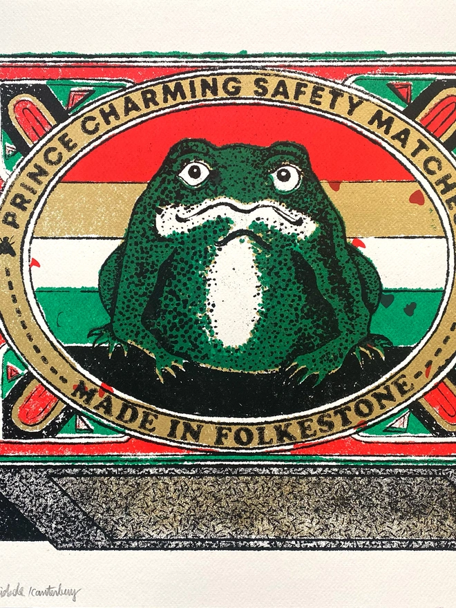  An image of a limited edition A2 screenprint. The print depicts a retro-style matchbox illustration with a central image of a green frog with a white belly, set against a background of horizontal red, white, and green stripes. The frog is framed by a gold-bordered oval. Text at the top of the oval reads "PRINCE CHARMING SAFETY MATCHES," and at the bottom, "MADE IN FOLKESTONE." The matchbox border is red with ornate details and a green trim. The match tips on the left are a textured gold. Below the main image, the print is signed and numbered 5/11. The entire image has a grainy texture, characteristic of a vintage screenprint.