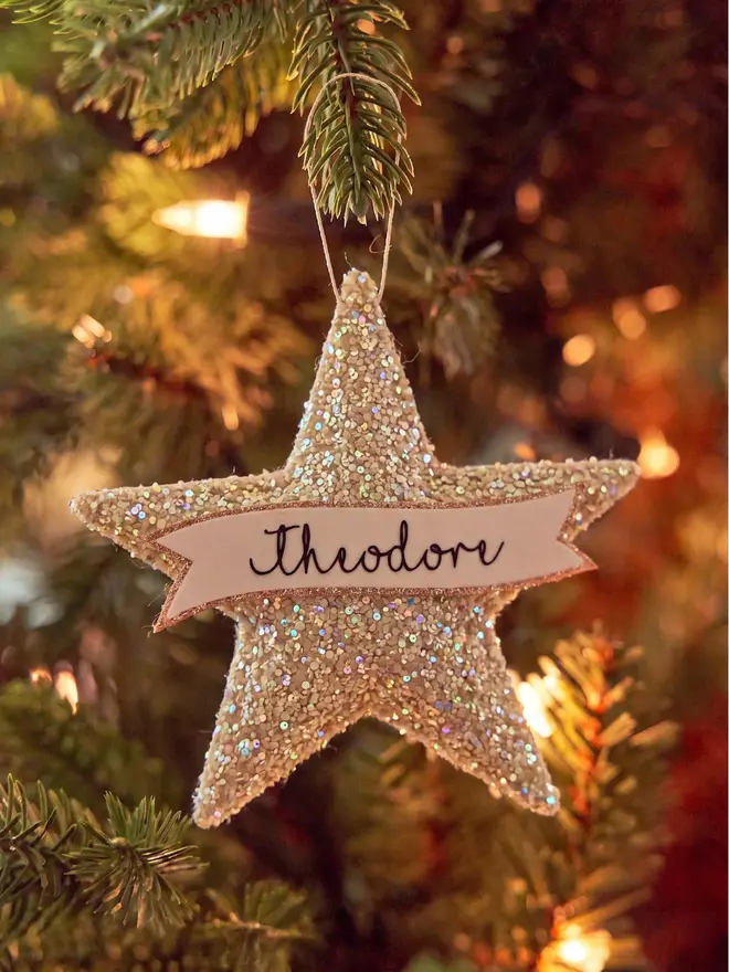 Beige glittery star decoration hung on a Christmas tree, personalised with the name 'Theodore' stitched onto the front in black on a white banner