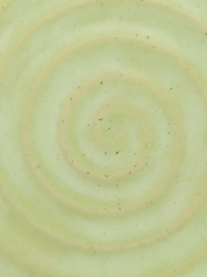 A close-up of a handmade ceramic glaze swatch in soft spring green, showing a smooth concentric swirl texture with a delicate, even tone throughout