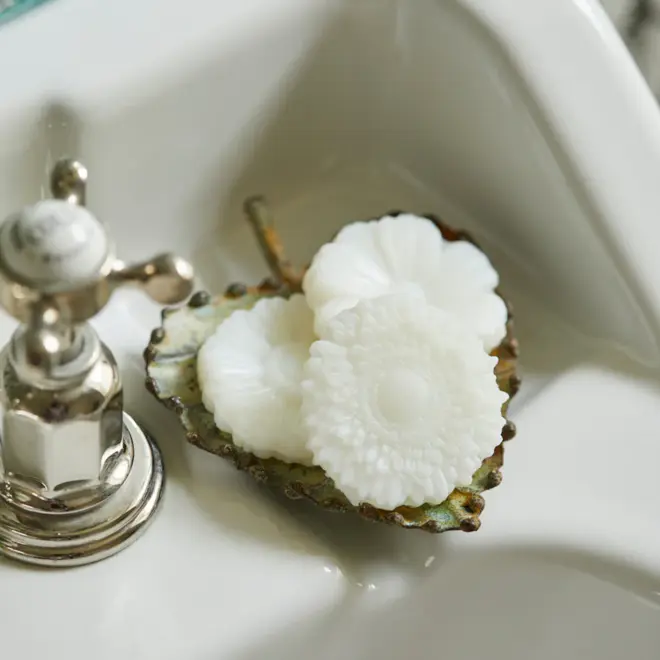 three white, flower shaped soaps sitting in a dark gold, leaf shaped dish, rest on a white sink