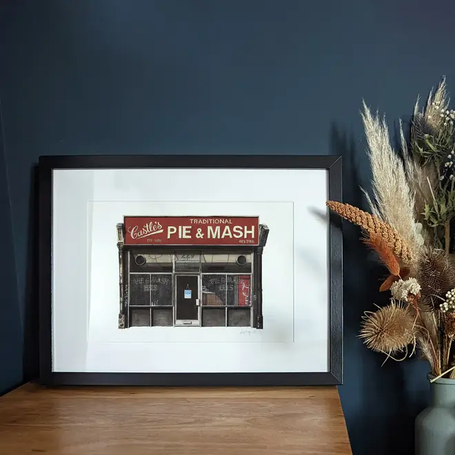 Camden Castle's Pie & Mash Shop Giclée Print (Unframed), a print of a shop sitting in a frame on a wooden surface against a navy blue backdrop. It is accompanied by some flowers. 