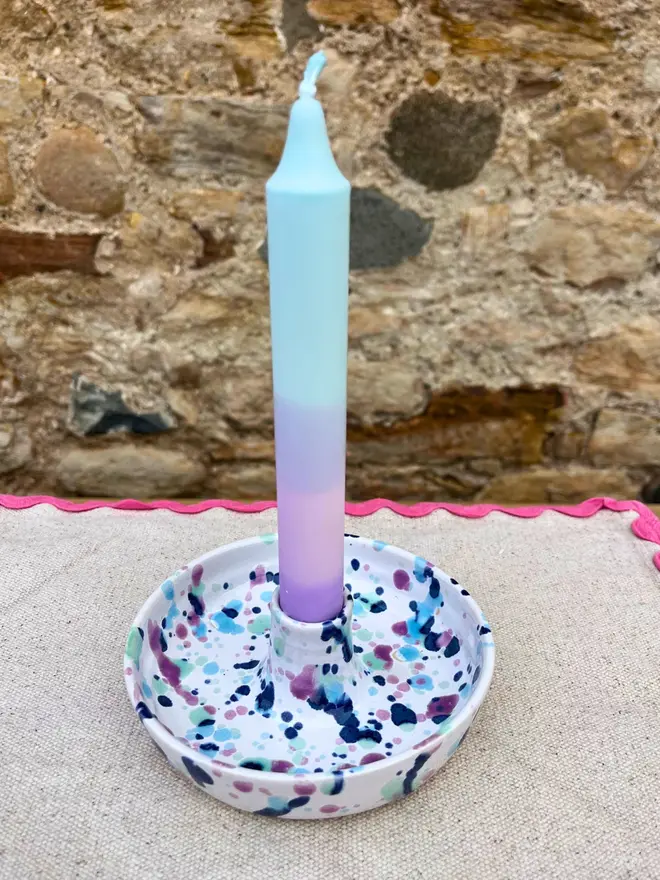 Sweetpea Candle Stick Holder, a colourful candle stick holder sitting on a stone surface against a brick backdrop. It is accompanied by a blue candle stick. 