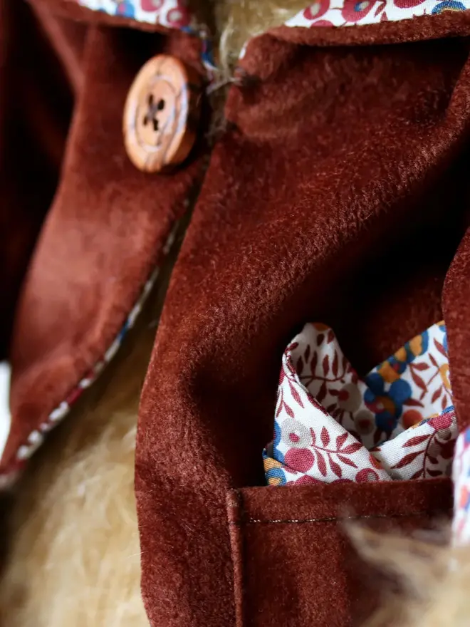 Brown teddy bear wearing a burgundy cloak with flower details and a brown button, sitting upright against a white background.