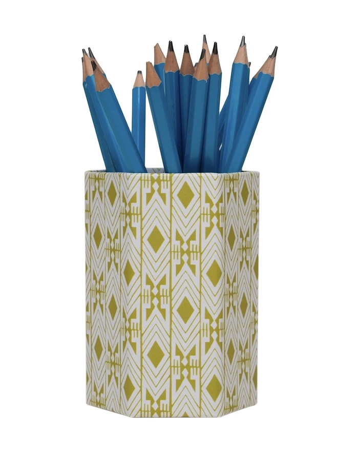 Tijan Hexagonal Brush Pot, a colourful patterned pencil pot sitting on a white surface against a plain backdrop. It is filled by some pencils. 