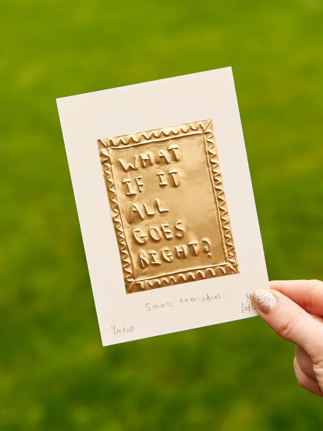 'What If It All Goes Right?' Gold Tin Embossing with a white border, held up in the garden 