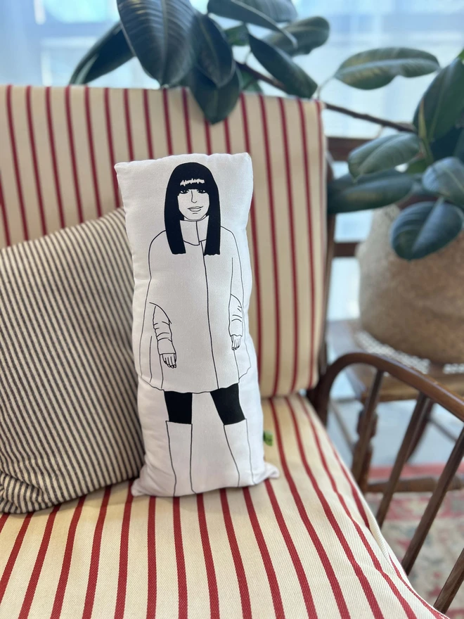 Black and white illustrated screen printed cushion of Traitors host Claudia Winkleman. The cushion sits on a striped sofa.