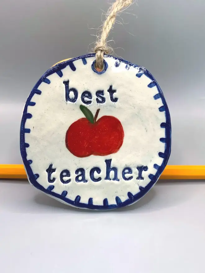 'Best Teacher' Handcrafted Ceramic Ornament, a ceramic apple ornament sitting on a plain white surface hanging from a pencil. 