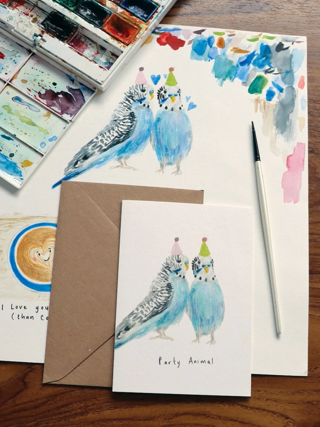  Desk Shot Of The Party Budgies Greeting Card Sitting Along Side The Original Hand Painted Watercolour Illustration, Paintbrush and Palette