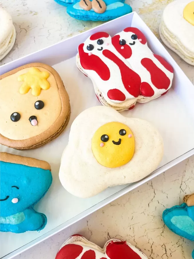 a box filled with macarons decorated to look like cartoon breakfast food characters - fried egg, toast, bacon rashers & coffee cup