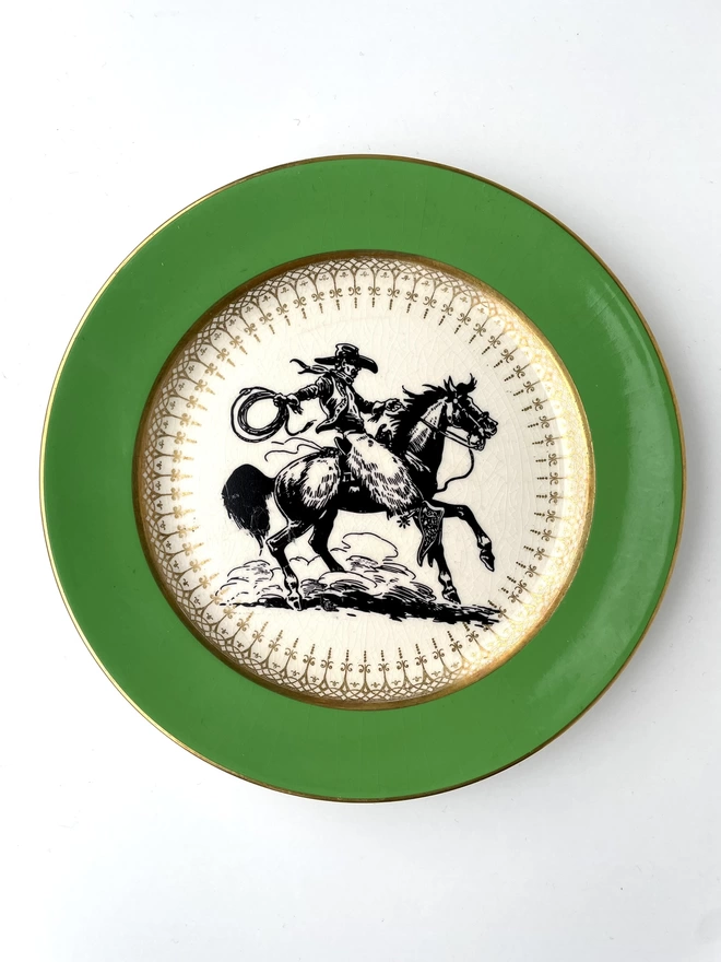 vintage plate hanging on a white wall, it has a fine gold edge and green border, with a printed vintage illustration of a cowboy in the middle 