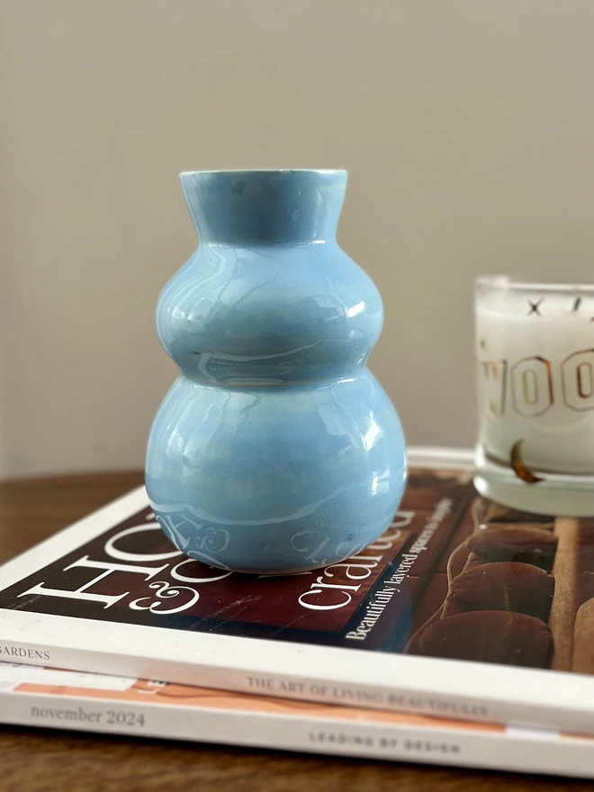 sky blue chubby bud vase sitting on a stack of magazines on a coffee table with a candle in the background. The bud vase has curvy side with a flared rim 