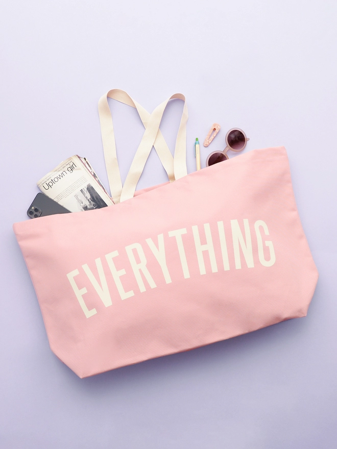 Everything oversized canvas tote bag in Pink held laying on a purple background with accessories spilling out