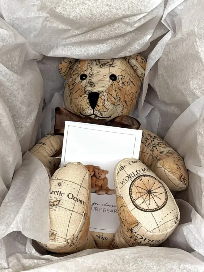 Teddy bear made from map-print fabric lying in a box, wearing a brown bow.