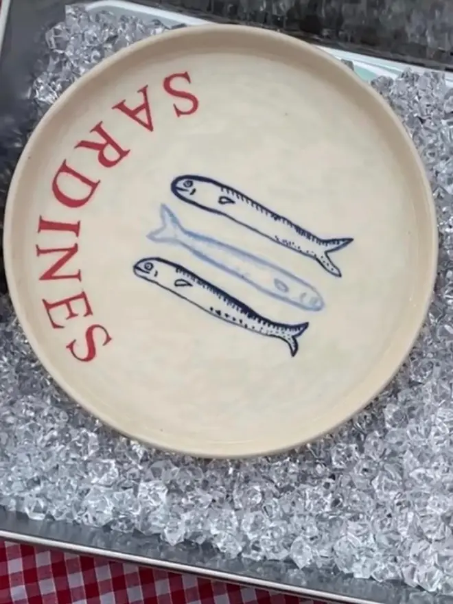 'Sardines' Red Wording Ceramic Side Plate With 3 Fish, a ceramic side plate sitting on ice with a fish design. 