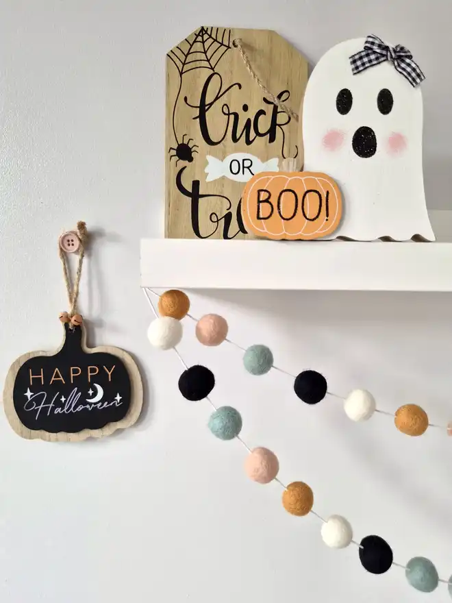 A wooden Halloween ghost on a shelf, with a felt ball garland hanging underneath in  boho colours.