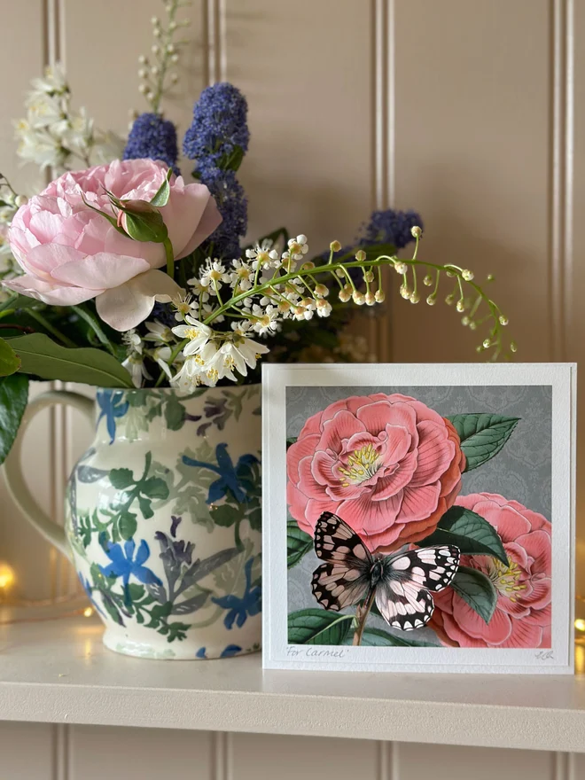 Mother’s day card, showing with a beautiful, pink Camellia flowers with a white butterfly. The card can be personalised on the bottom left corner. The camellias are against a stylish grey background. Sitting on the flower is a white butterfly. The butterfly is 3 dimensional and the wings can be folded open for display. The card is sitting on a dresser displayed in the home setting.