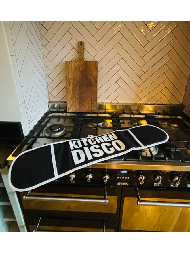 Black oven glove with white text that says KITCHEN DISCO laying on hob with tiles and chopping bard in the background