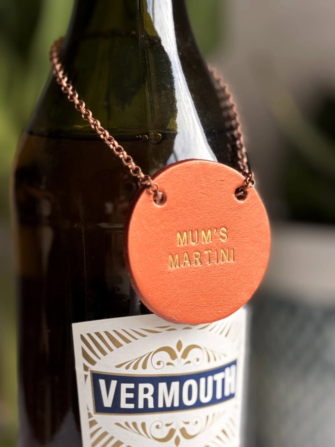 A dark glass bottle with a copper leather tag hanging around the neck by a delicate chain. The tag is hand-stamped with 'MUM'S MARTINI' in gold lettering, with pink tulips and a rustic background. A perfect personalised Mother’s Day gift or home bar accessory