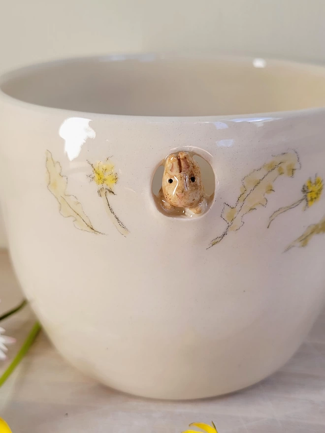 a beige pottery bunny looking out of a cut-out in a hand thrown ceramic beaker with hand painted flowers and leaves