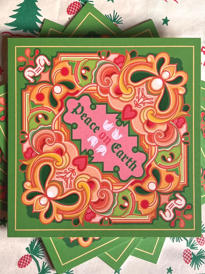 Three square cards displayed on top of each other, sitting on Christmas wrapping paper. An abstract Christmas card design with Peace on Earth at the centre, surrounded by a multi-coloured design on a green background. The card features greens, pinks and oranges.