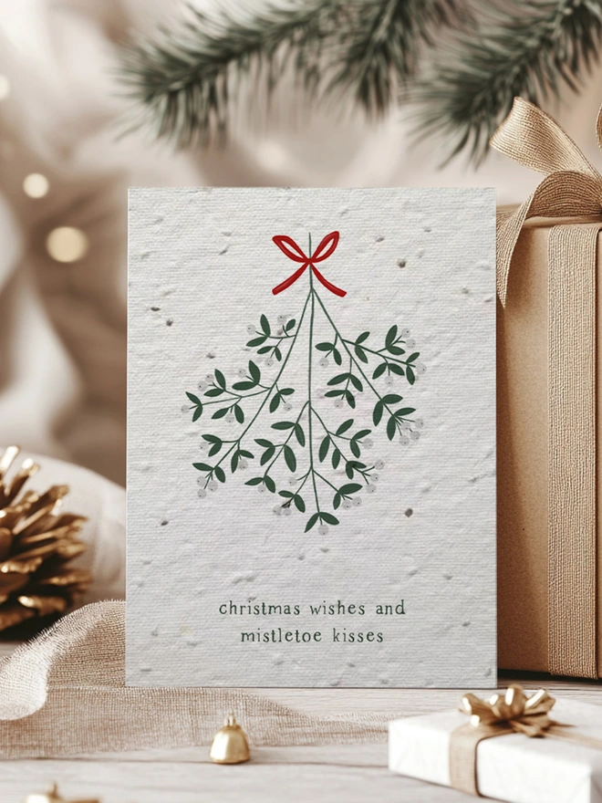 Christmas Card with illustration of mistletoe with 'Christmas Wishes and Mistletoe Kisses'. In the background is Christmas presents, greenery and pine cones.
