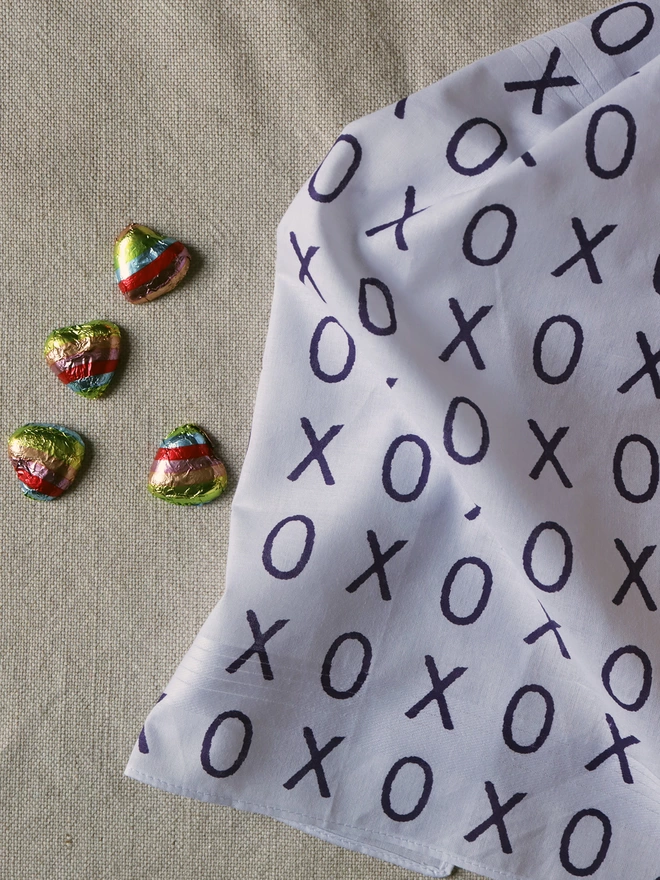 A Mr.PS Hugs and Kisses Handkerchief printed in deep violet laid on a linen tablecloth with some foil-wrapped chocolate hearts