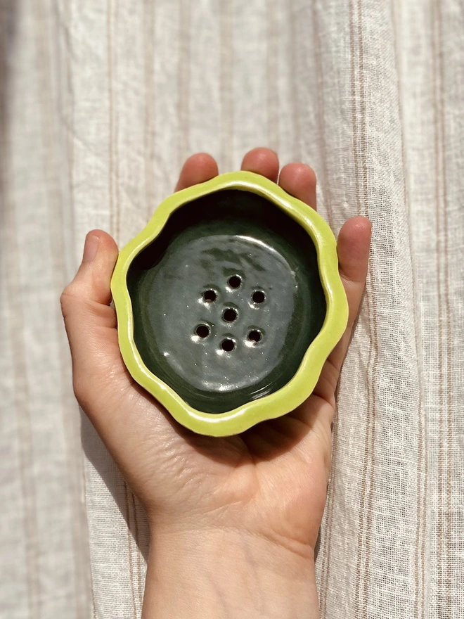wavy rimmed soap dish held in the palm of a hand against a fabric background. the soap dish is lime green on the outside and dark green on the inside
