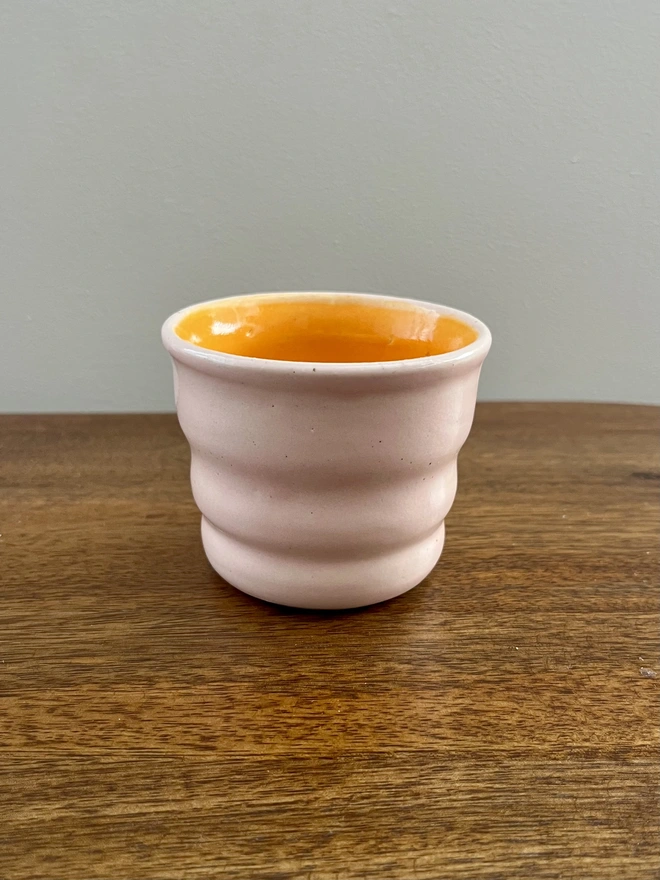 mini ceramic tumbler with wavy sides on a wooden table. the tumbler has a blush pink glaze on the outside and tangerine orange on the inside and rim. 