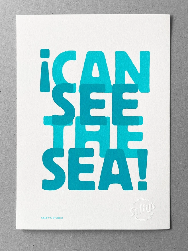Full view of the A4 I Can See the Sea print. Two colours turquoise and teal, slightly overlapping words evoking the sense of waves. The print sits on a greyboard background.