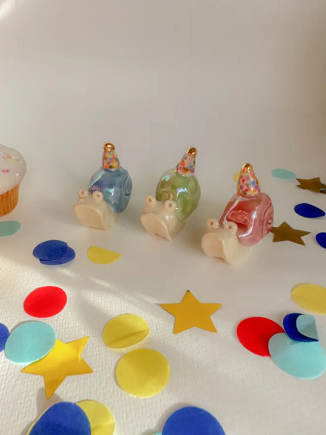 Handmade Porcelain Snail Cake Topper With Golden Hat, grouped together on a table with party decorations. 