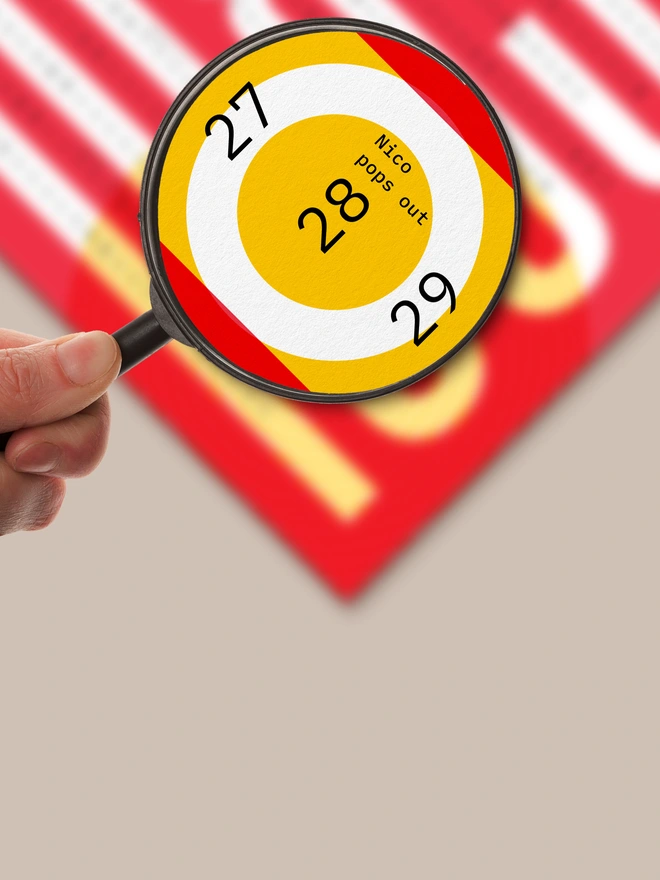 Hand with magnifying glass, focus on a printed poster in red and yellow with highlighted date "28" and text "Nico pops out"